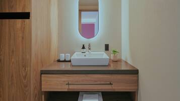 Une salle de bain moderne avec un meuble-lavabo en bois, un lavabo blanc, un miroir avec un cadre rose et une petite plante en pot.