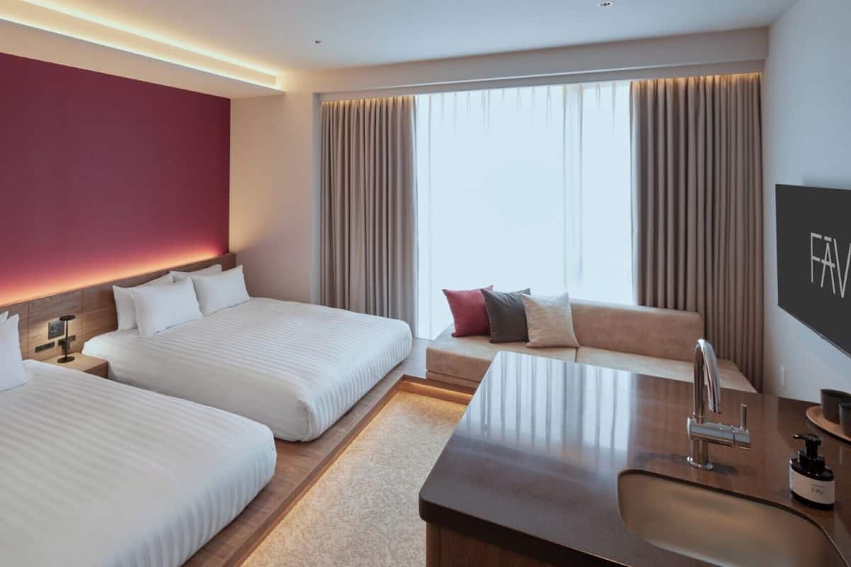 A hotel room with two beds, a sofa, a TV, and a bathroom with a mirror and sink.