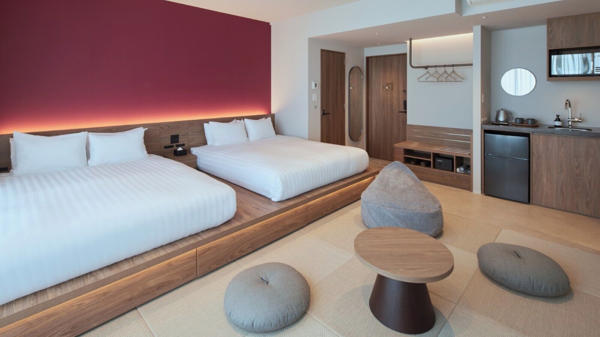 A modern hotel room with two beds, a small kitchenette, a seating area with ottomans, and a red accent wall.