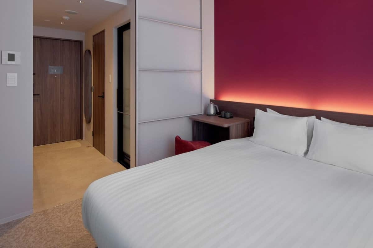 A hotel room with a large bed, a nightstand, a red pillow, and a wall with a red accent.