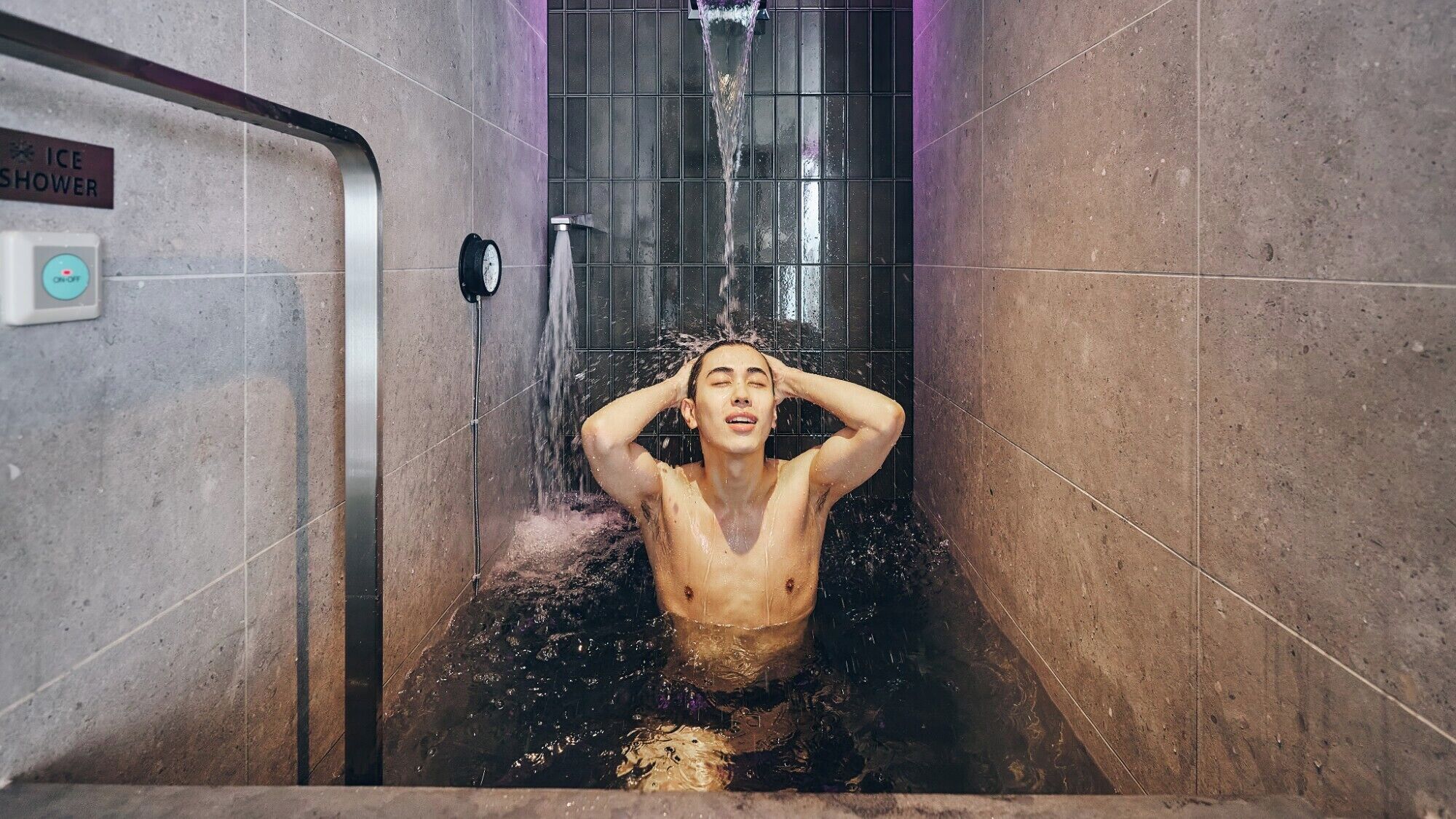 Une personne dans une baignoire où l’eau coule, un panneau indiquant une douche froide, et un panneau de commande avec un bouton d’alimentation.