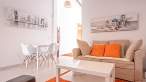Una sala de estar moderna con un sofá blanco, cojines naranjas, una mesa de centro blanca y una alfombra roja. Hay dos sillas blancas y una mesita con una lámpara.