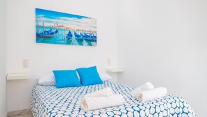 Habitación de hotel con una cama que tiene una colcha de diseño azul y blanco, dos almohadas azules y dos toallas blancas enrolladas. En la pared sobre la cama, hay un cuadro con barcos en el agua.