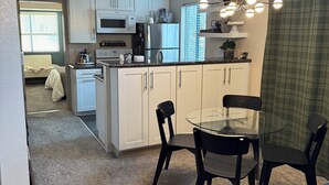 Cocina compacta con gabinetes blancos, refrigerador, microondas y comedor con mesa de vidrio y sillas negras.