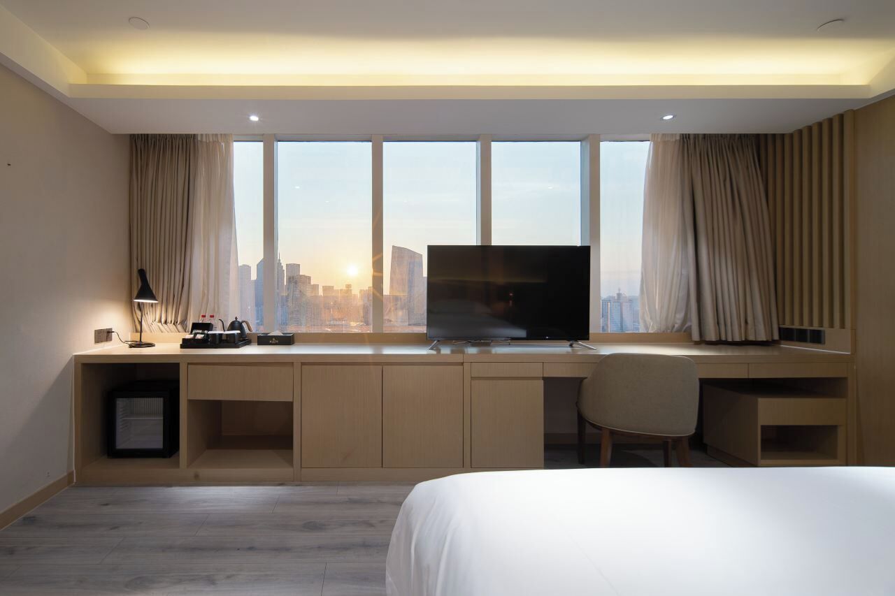 Habitación de hotel con una cama grande, un escritorio con televisor y vistas a un paisaje urbano a través de amplios ventanales.