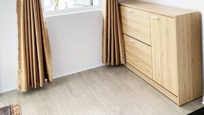 Una habitación con cortinas color beige atadas en la parte superior, un armario de madera y una ventana con vistas a edificios y follaje.