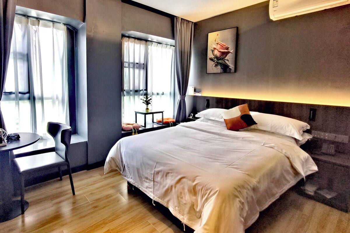 Ein modernes Hotelzimmer mit einem großen Bett, einem Schreibtisch und einem Sessel. Ein Fenster mit Vorhängen, ein Blumentopf und ein Bild an der Wand sind ebenfalls vorhanden.