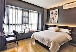 Habitación de hotel moderna con una cama grande, un escritorio con silla, una mesita con una planta y una ventana con cortinas.
