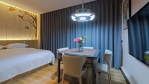 Una camera d'albergo con un letto, una scrivania con sedie, un tavolo con un vaso di fiori, una TV a muro e un lampadario.