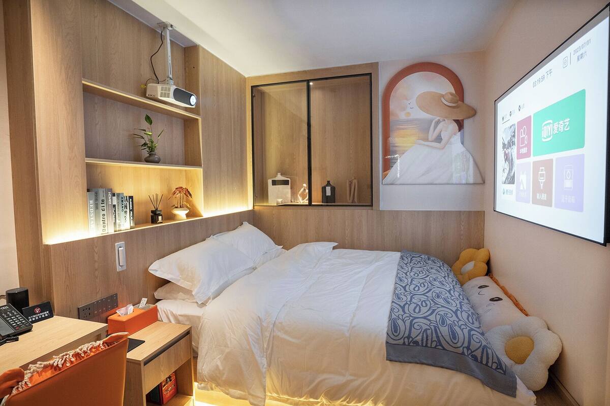 Ein modernes Hotelzimmer mit einem Bett, einem Schreibtisch mit Computer, einem Bücherregal und einem an der Wand befestigten Projektor.