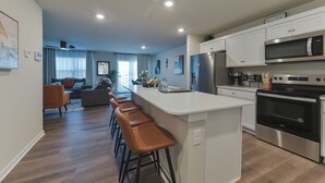 A modern kitchen with a central island, bar stools, stainless steel appliances, and a dining area in the background.
