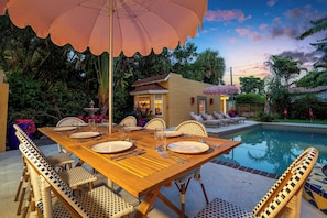 An outdoor dining area with a wooden table set for six, surrounded by wicker chairs, under a large umbrella.