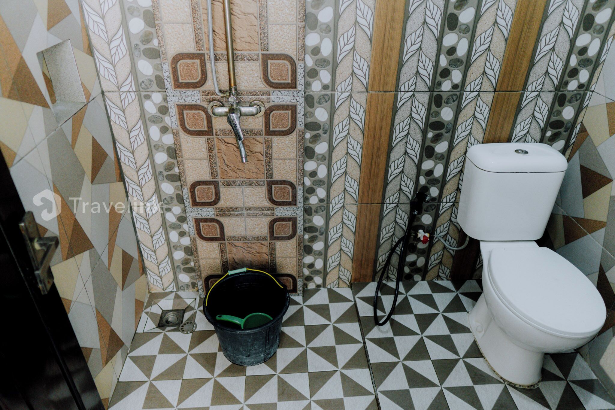 A bathroom with a geometric tile floor, a toilet, a shower with a faucet, and a bucket.