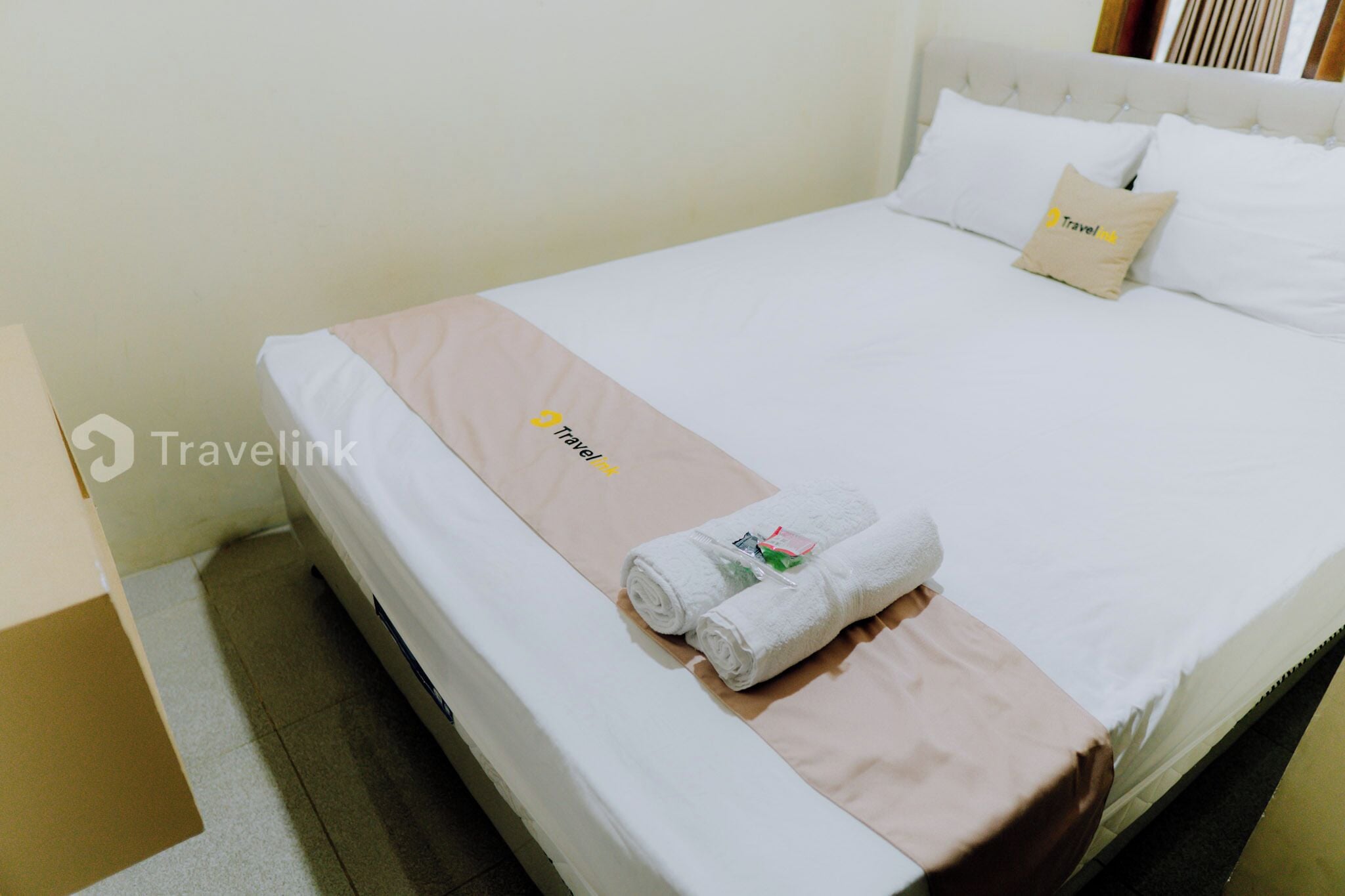 Una cama bien tendida con una toalla con el logotipo de Travellink y un almohadón beige.