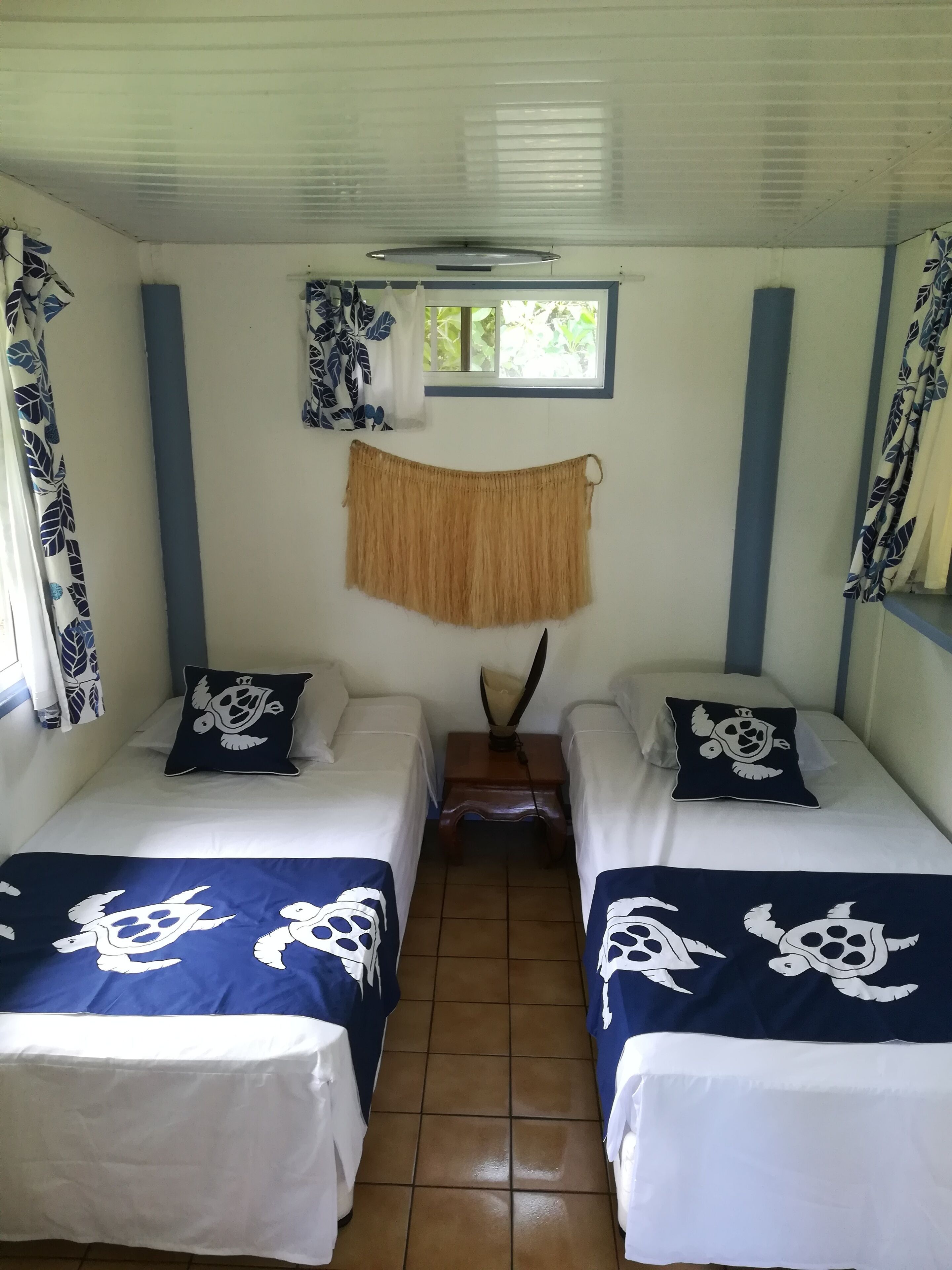 Una habitación con dos camas, cada una con una colcha blanca y azul con diseño de tortuga. Hay una ventana con marco azul y una cortina con estampado de hojas.