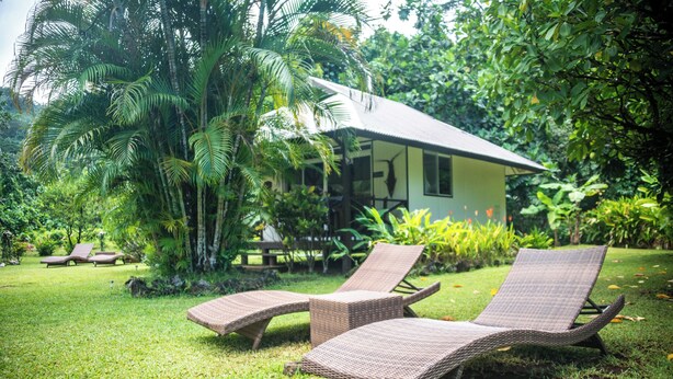 Een tropische villa met rieten dak, omgeven door weelderige begroeiing en twee rieten loungestoelen.