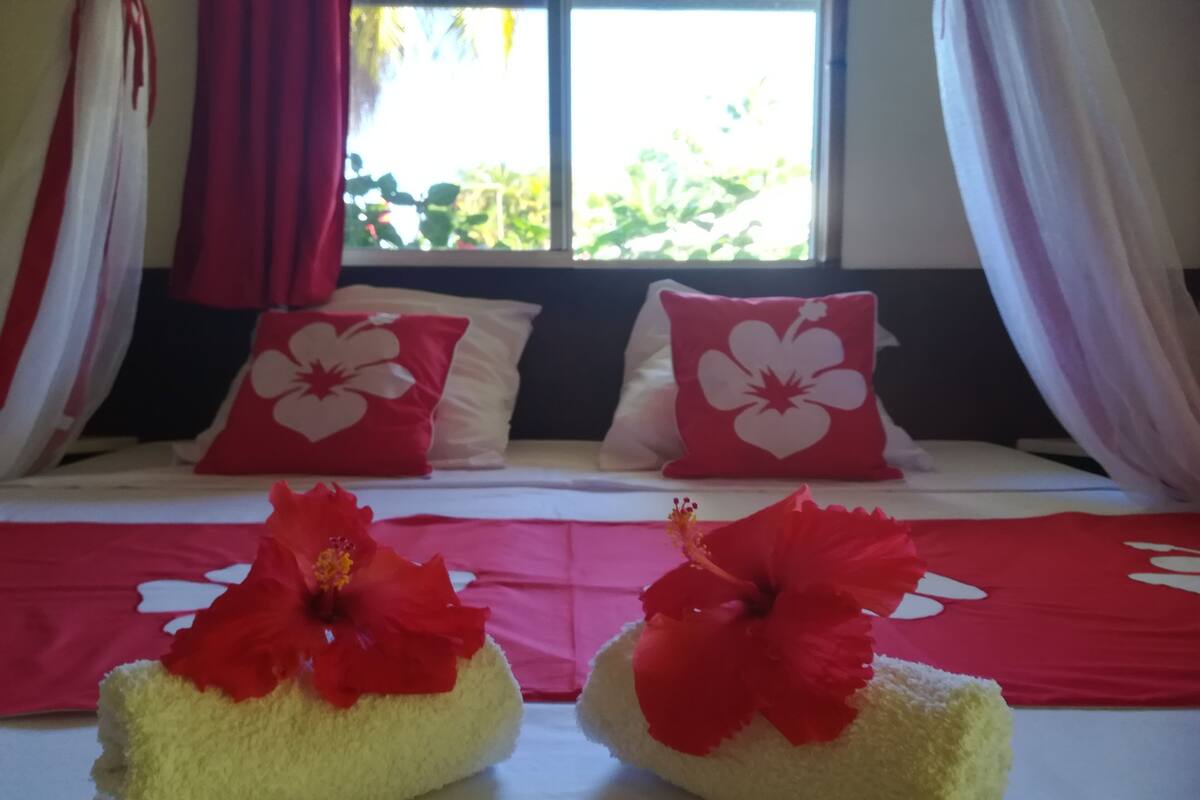 A hotel room with a bed, two red pillows with white floral designs, and a red and white bedspread.