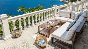 A balcony with a view of the sea, a wooden table, a wicker basket, and a sofa with cushions. - Villa Linda (Blato)