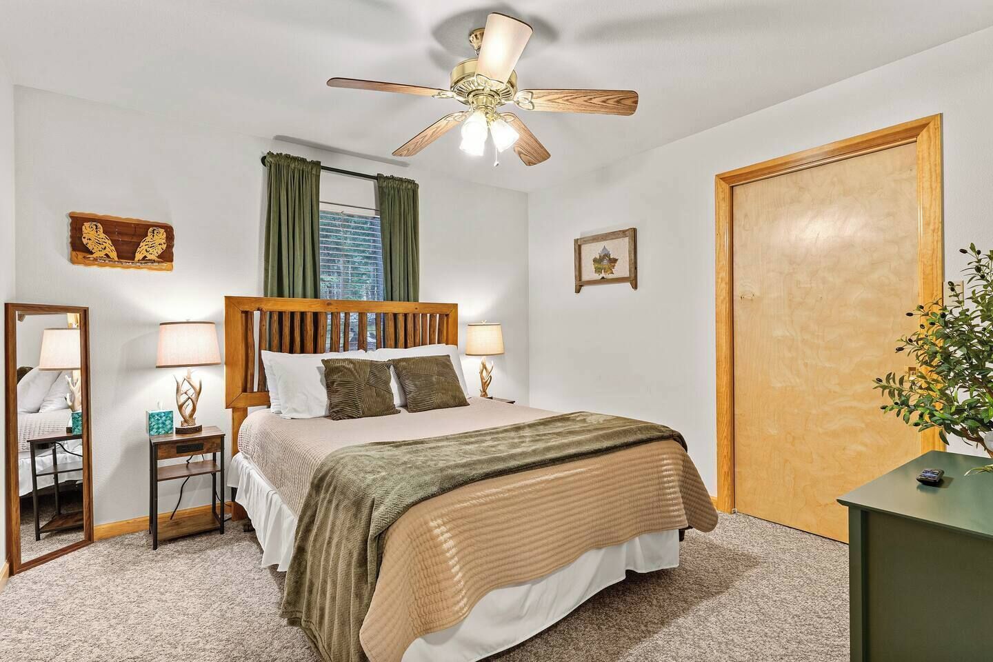 A hotel room with a bed, a ceiling fan, a window with curtains, a wooden door, and a small table with a plant.