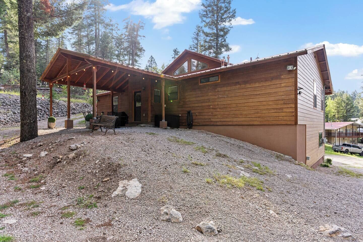 A wooden cabin with a covered patio area, a gravel driveway, and a view of the surrounding forest.