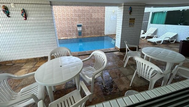 Un área de piscina interior con paredes de azulejos, una piscina y sillas y mesas de plástico blanco.