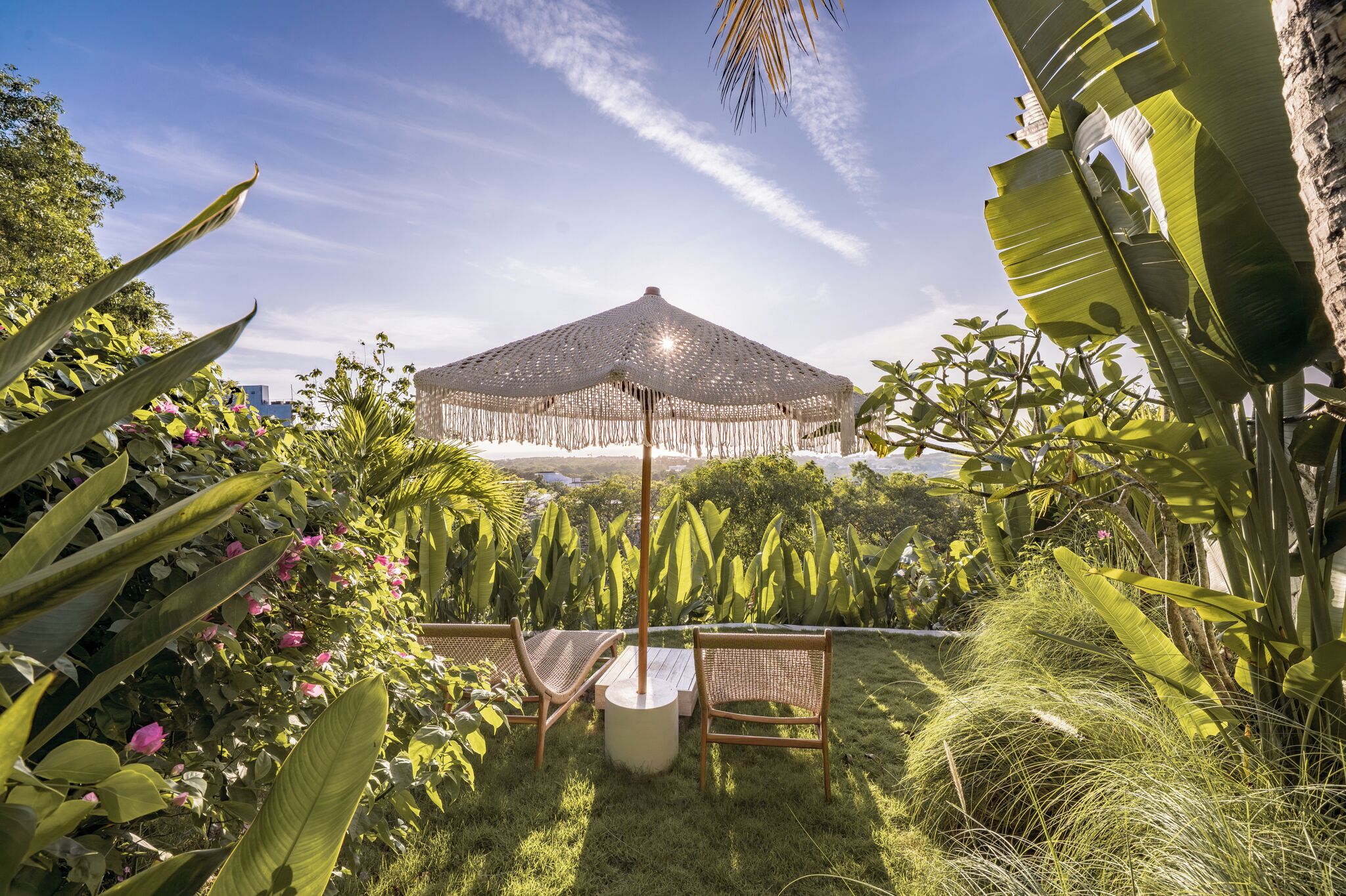 A garden with a thatched umbrella, wicker chairs, and lush greenery.