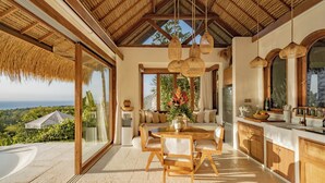 A spacious room with a large dining table, wooden chairs, a kitchen area, and a view of the ocean. - Munay Wasi Uluwatu (Pecatu)