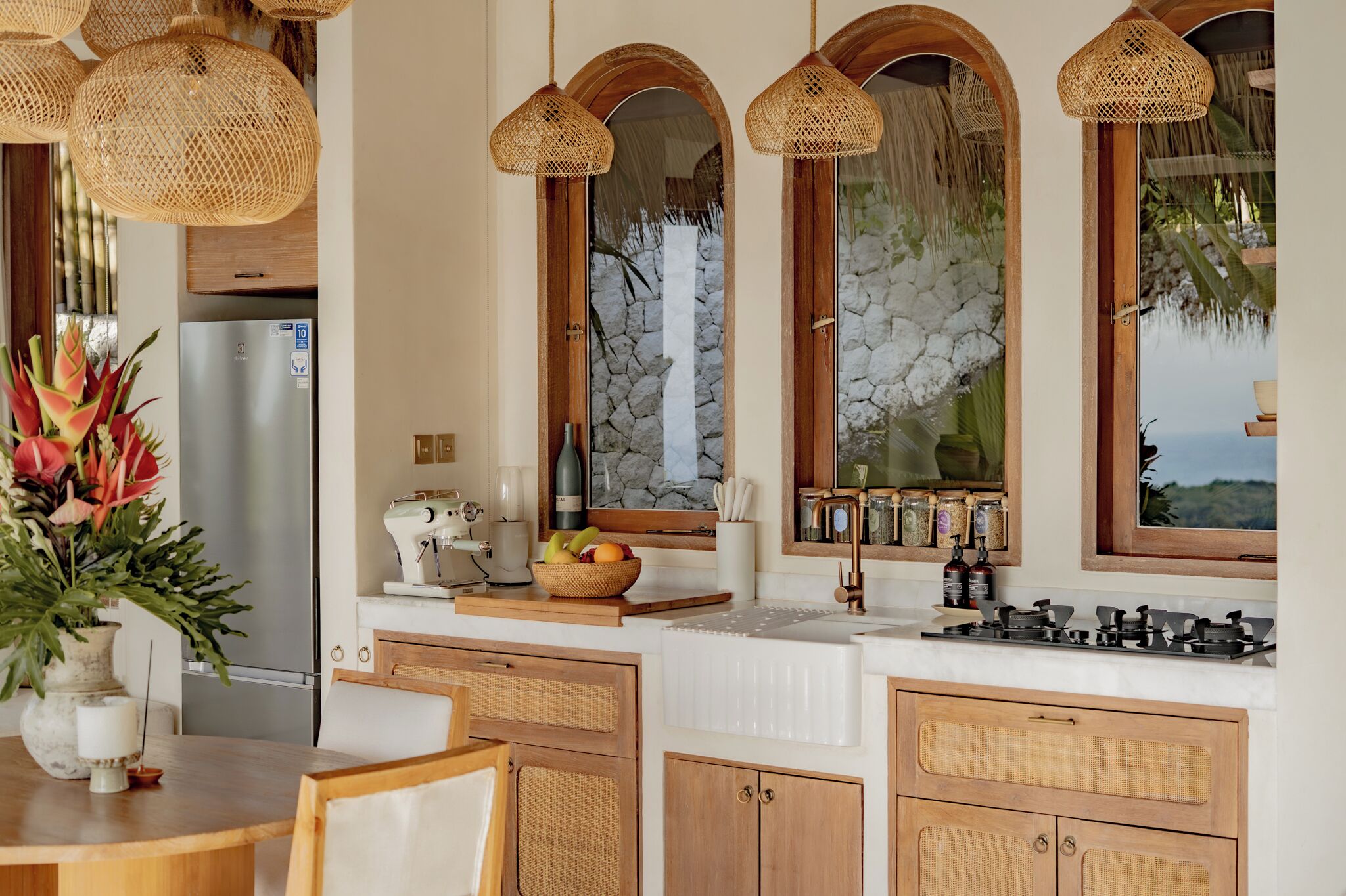 A kitchen with a refrigerator, a sink, a stove, wicker hanging lights, and a stone wall.