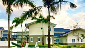 A grassy area with a seesaw and a small blue table, surrounded by palm trees and a building with a blue roof.