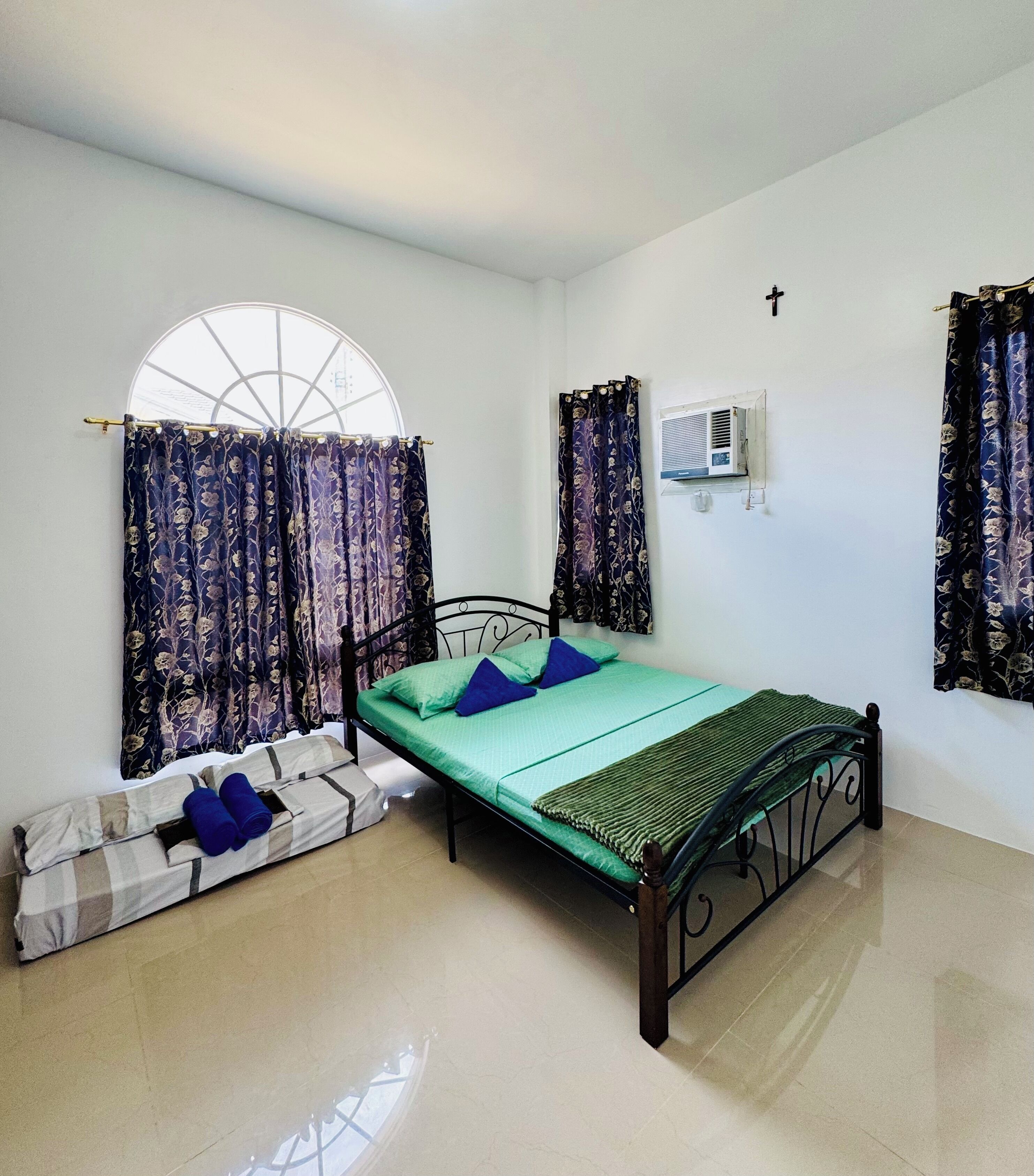 Un lit simple avec un matelas vert, deux oreillers bleus et une couverture à carreaux. Il y a une fenêtre avec un rideau, un climatiseur fixé au mur et une croix accrochée au mur.