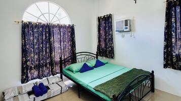 Un lit simple avec un matelas vert, deux oreillers bleus et une couverture à carreaux. Il y a une fenêtre avec un rideau, un climatiseur fixé au mur et une croix accrochée au mur.