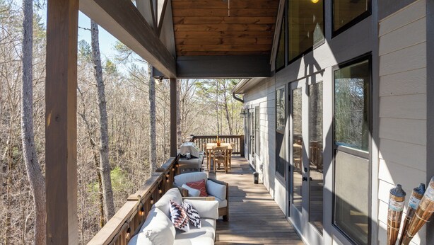 Una terraza techada al aire libre con mobiliario de madera, ventanas amplias y vistas al bosque circundante.