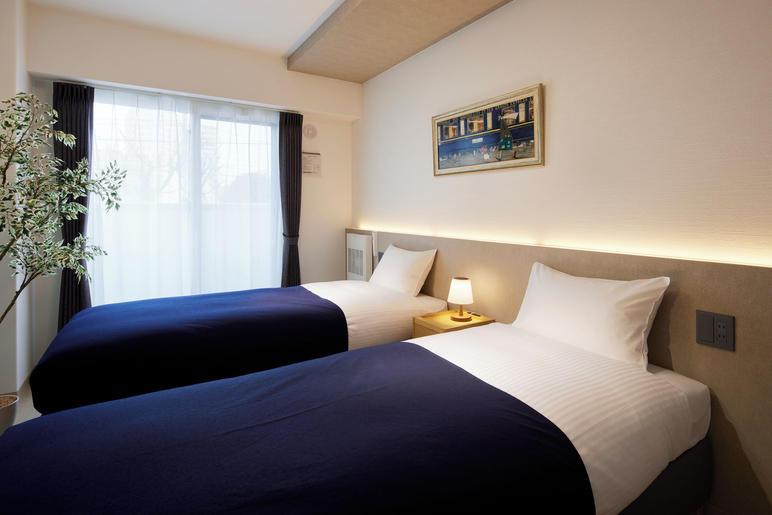 A hotel room with two beds, a nightstand with a lamp, a window with curtains, and a wall-mounted painting.