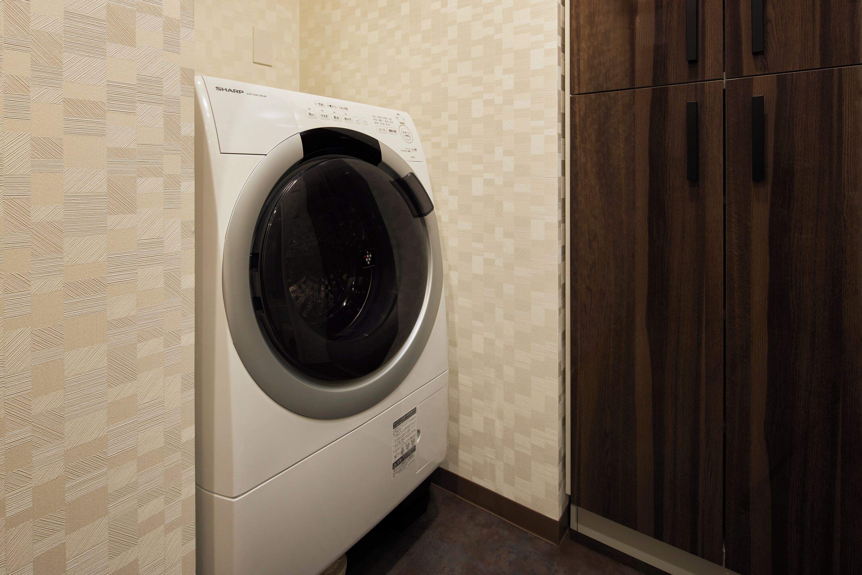 A compact, modern laundry room with a built-in washer and dryer, wooden cabinets, and patterned wallpaper.