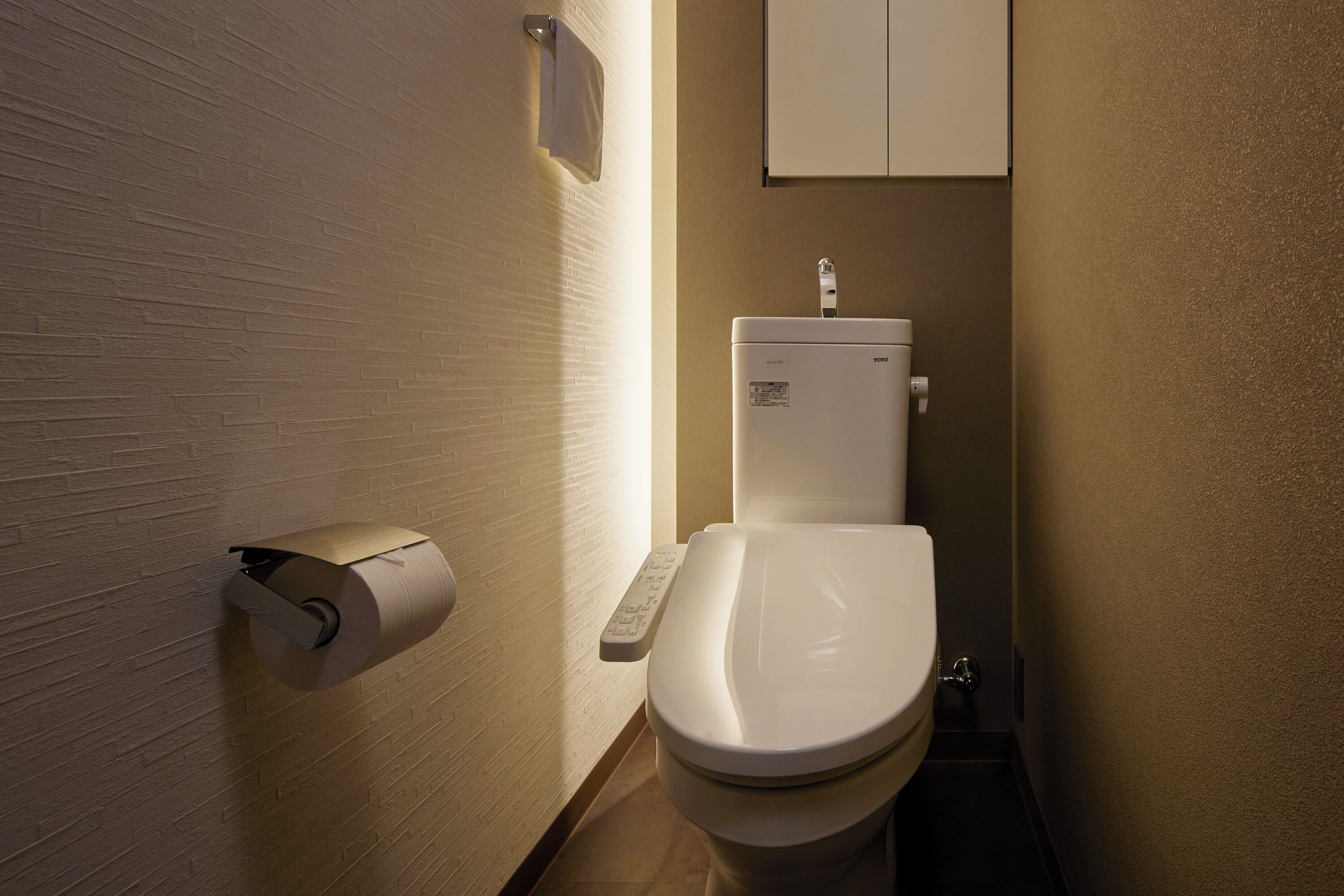Un baño moderno con inodoro, dispensador de toallas de papel y un mueble sobre el inodoro.