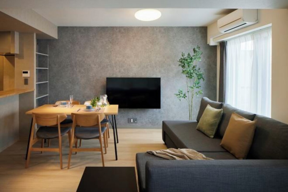 A modern living room with a flat-screen TV mounted on the wall, a sofa, a dining table with chairs, and a potted plant.