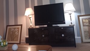 Olohuoneessa on seinällä tumma puinen tv-taso, jossa on litteänäyttöinen televisio, kaksi pöytävalaisinta ja kehystetty taideteos.