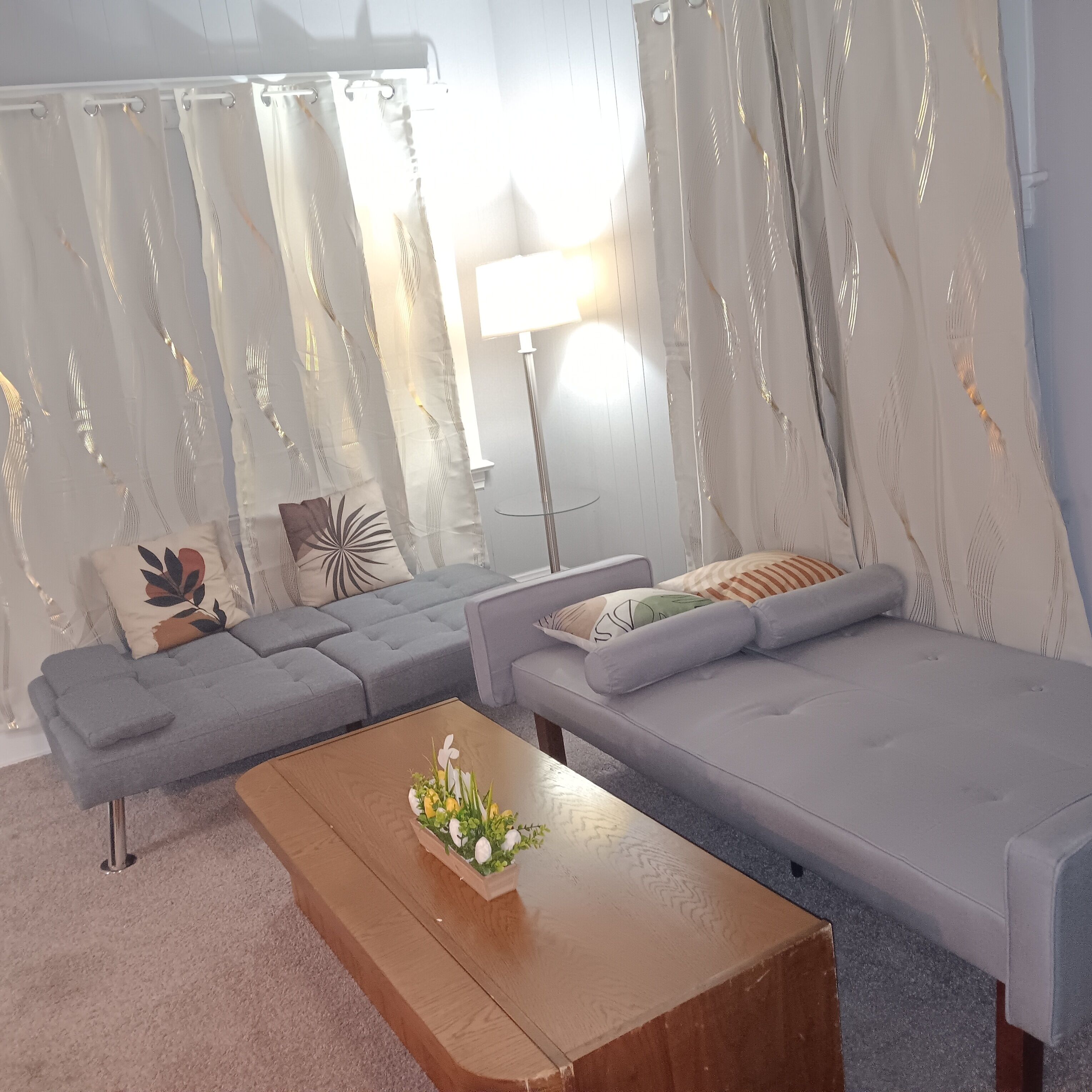 A modern living room with a grey sectional sofa, a wooden coffee table with a floral arrangement, and sheer curtains with a leaf pattern.