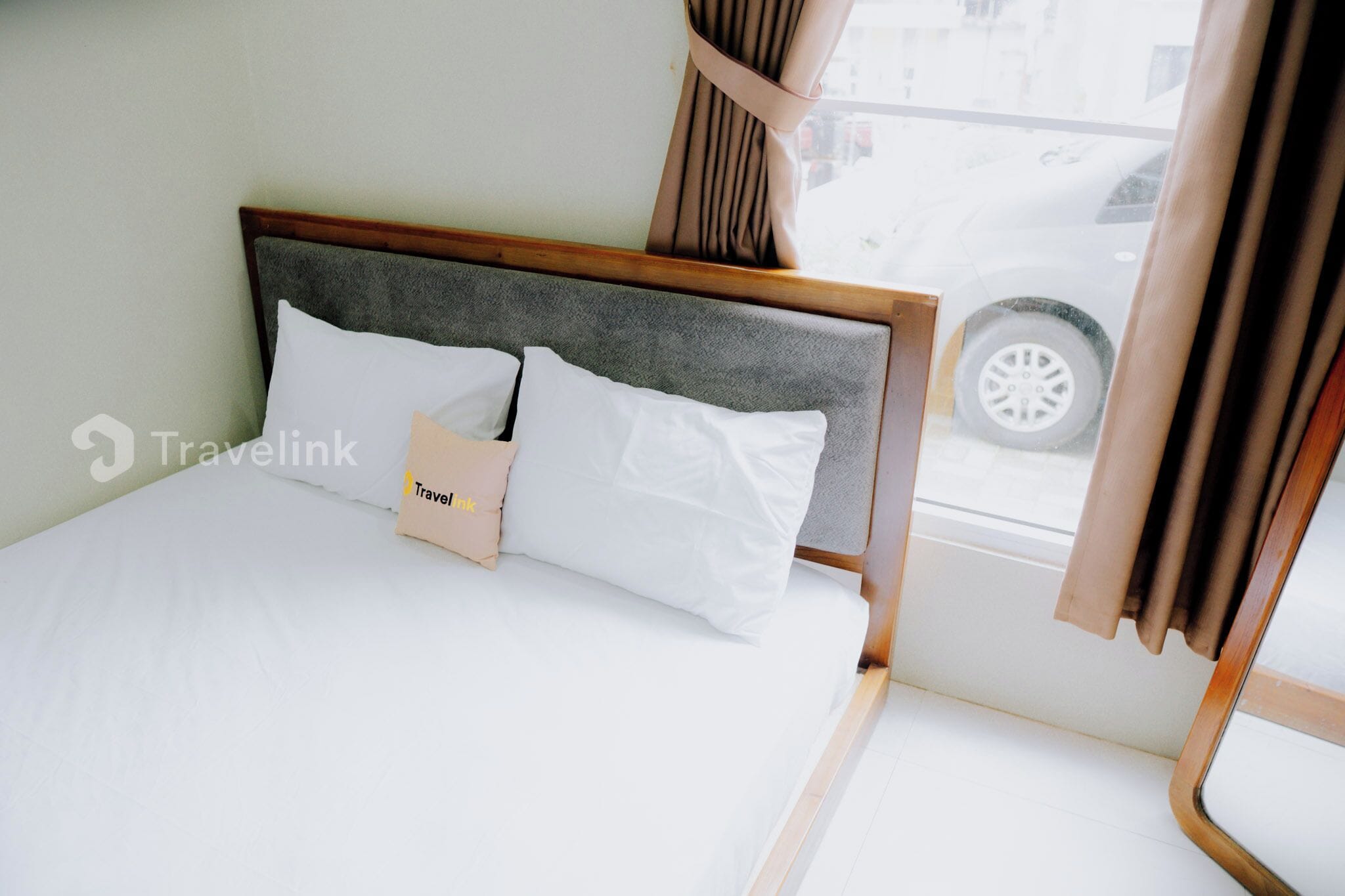 Una cama cuidadosamente tendida con un cabecero gris, ropa de cama blanca y una almohada de la marca Travelink.