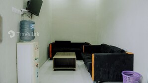 Een kleine, eenvoudig ingerichte kamer met een zwarte bank, een houten bijzettafeltje, een waterdispenser en een wandtelevisie.