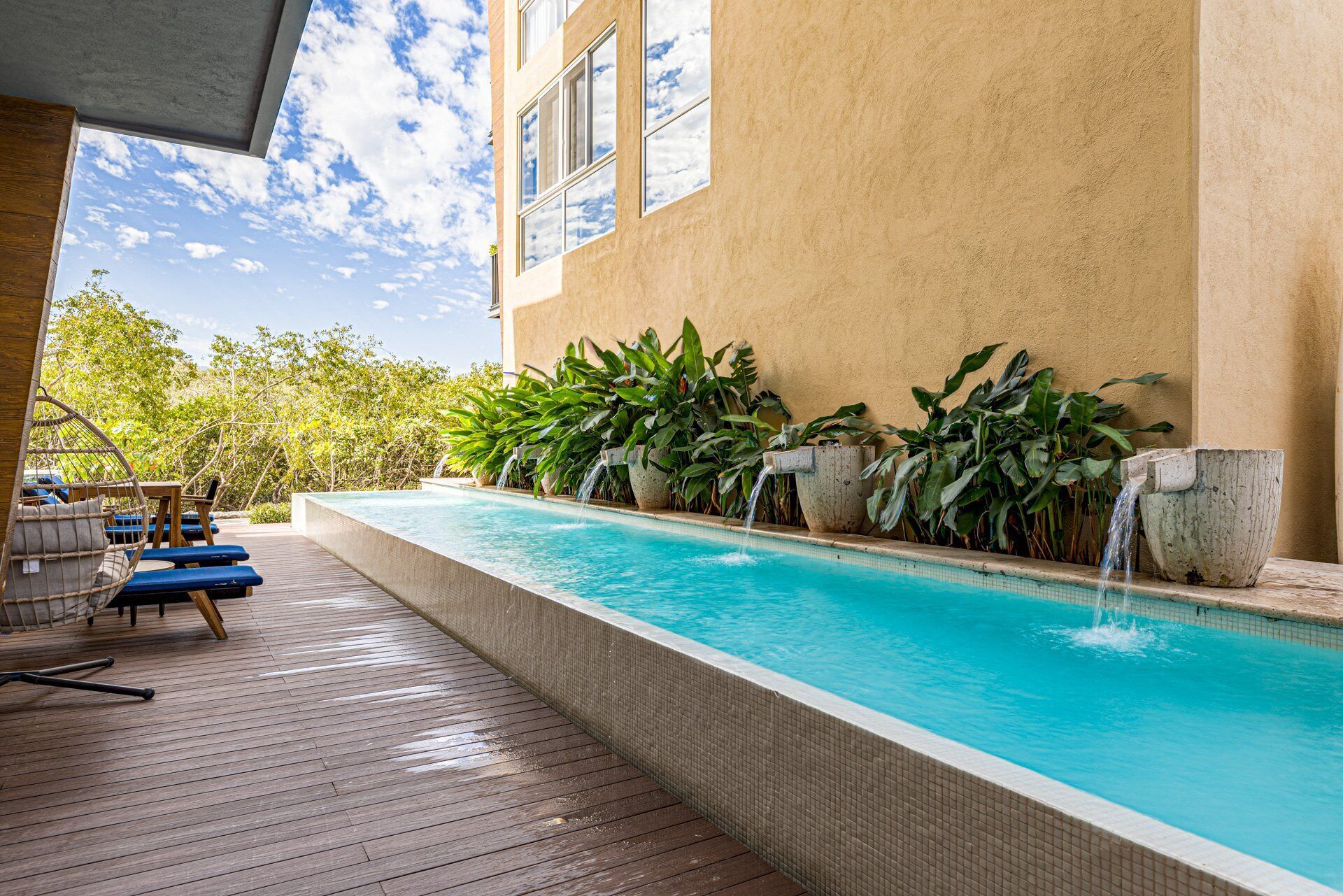 Une piscine dotée d’une terrasse en bois et de chaises longues, entourée de plantes tropicales et bordée d’un bâtiment aux grandes fenêtres.