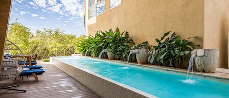 Une piscine dotée d’une terrasse en bois et de chaises longues, entourée de plantes tropicales et bordée d’un bâtiment aux grandes fenêtres.