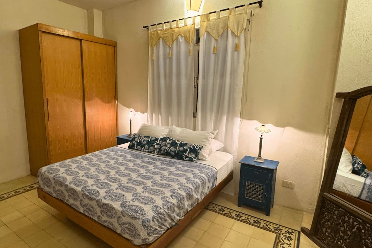 Un dormitorio con cama, mesita de noche, armario y ventana con cortinas.