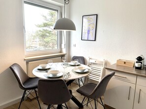 Ein Essbereich mit einem runden Tisch, gedeckt für vier Personen, Stühlen, einer Pendelleuchte, einem gerahmten Bild an der Wand und einer Küchenzeile mit einer Kaffeemaschine.