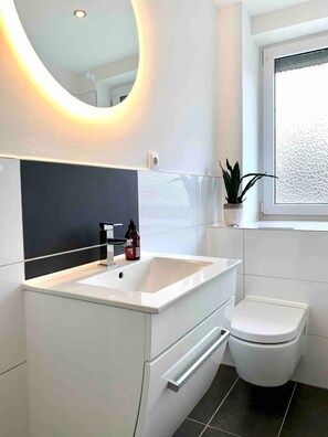 Ein modernes Badezimmer mit einem weißen Waschbecken, einer Toilette und einem Fenster mit Blick nach draußen.