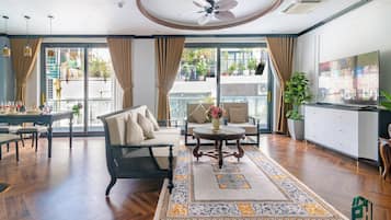 A spacious living room with a sofa, chairs, a coffee table, and a TV. There is a balcony with plants and a view of the outside.