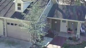 Une maison à deux étages avec un garage, une véranda et un petit arbre devant.