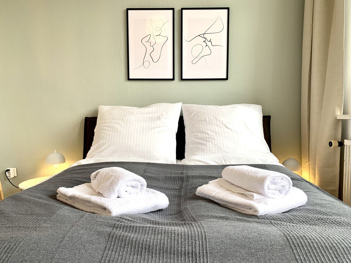 Een keurig opgemaakt bed met witte kussens en een grijze deken, met daarboven twee ingelijste kunstwerken en een bedlampje.