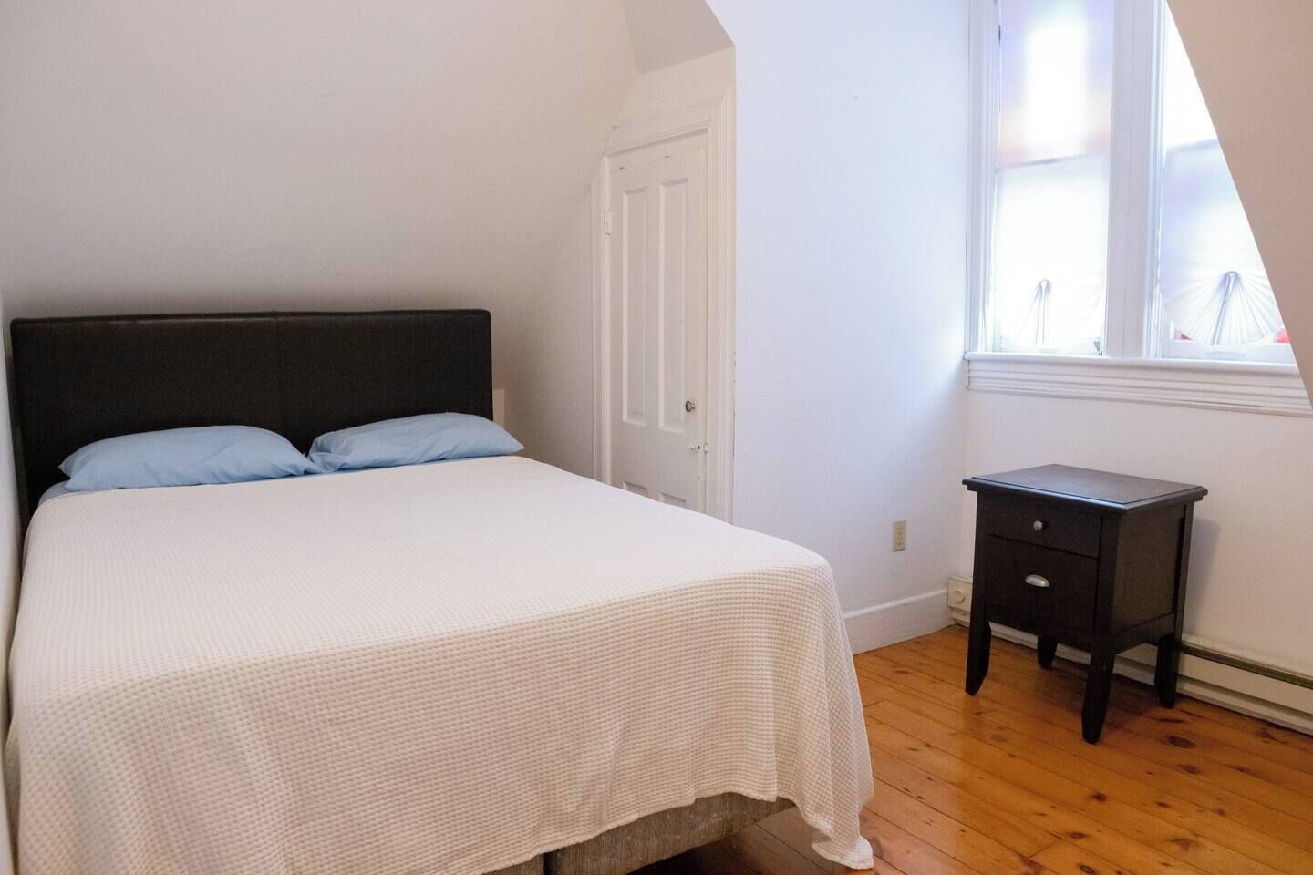 A bedroom with a bed, a nightstand, a door, and a window.