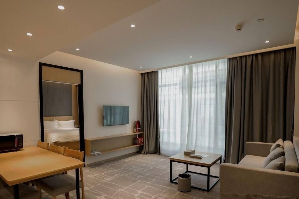 Ein modernes Hotelzimmer mit einem Bett, einer Sitzecke mit Tisch und Stühlen, einem Fernseher und einem großen Fenster mit Vorhängen.